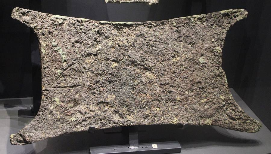 Lingote de cobre corroído con forma de piel de buey extendida, sobre un soporte de museo