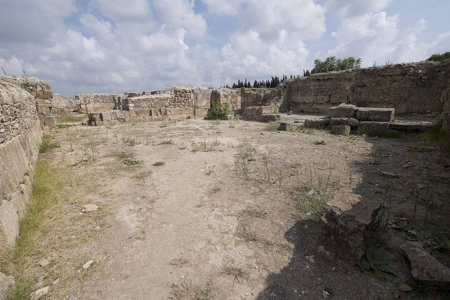 Explanada de ruinas con muros de sillares de piedra a media altura bajo un cielo nuboso